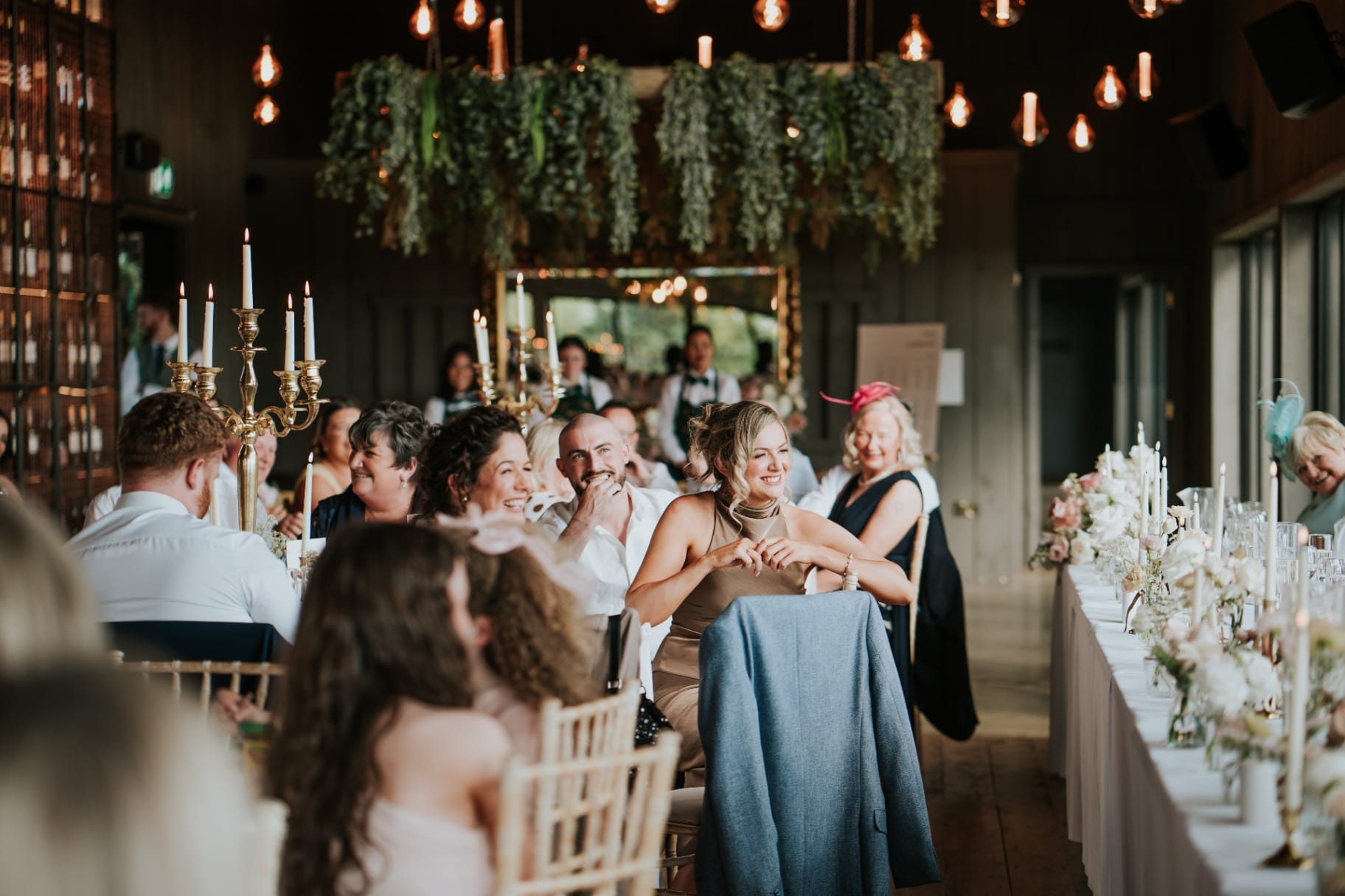 Wedding guests smiling during speeches beneath hanging greenery and warm lighting
