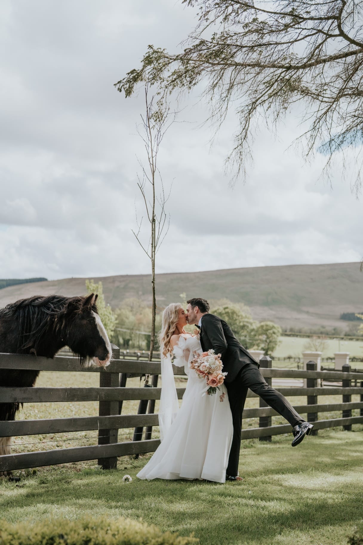 Groom dipping bride for a kiss beside the paddock with horse looking on