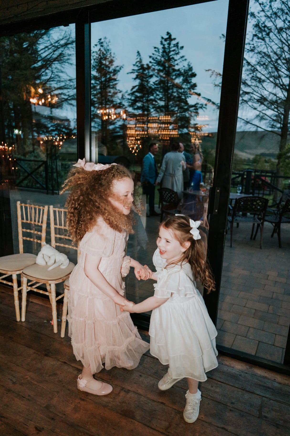 Two flower girls dancing together by the glass doors at dusk