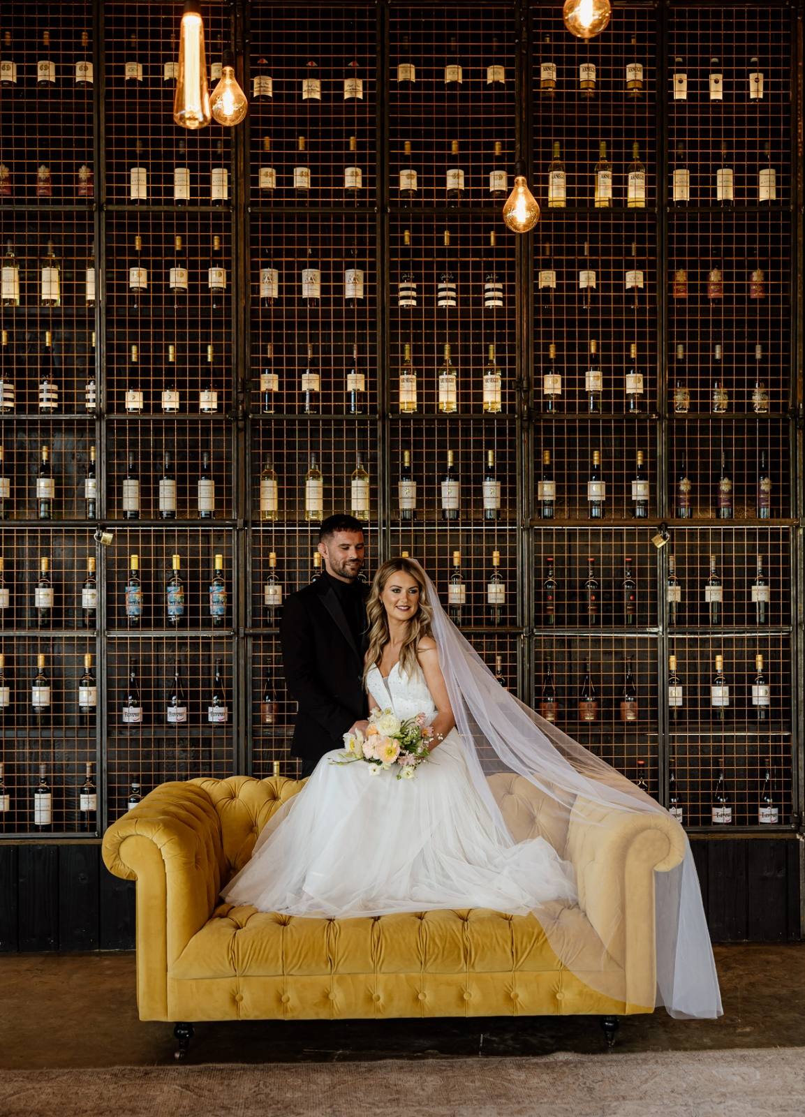Bride and groom seated on a mustard sofa in front of the Ballyness wine wall