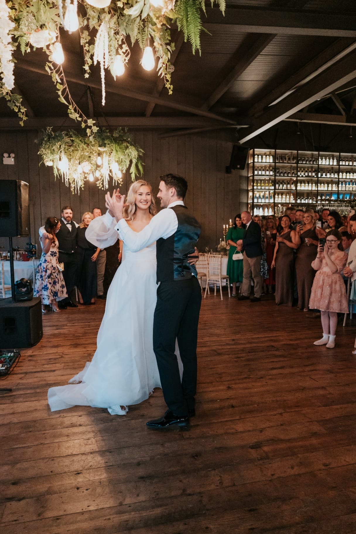 Newlyweds dance beneath botanical chandeliers as guests look on