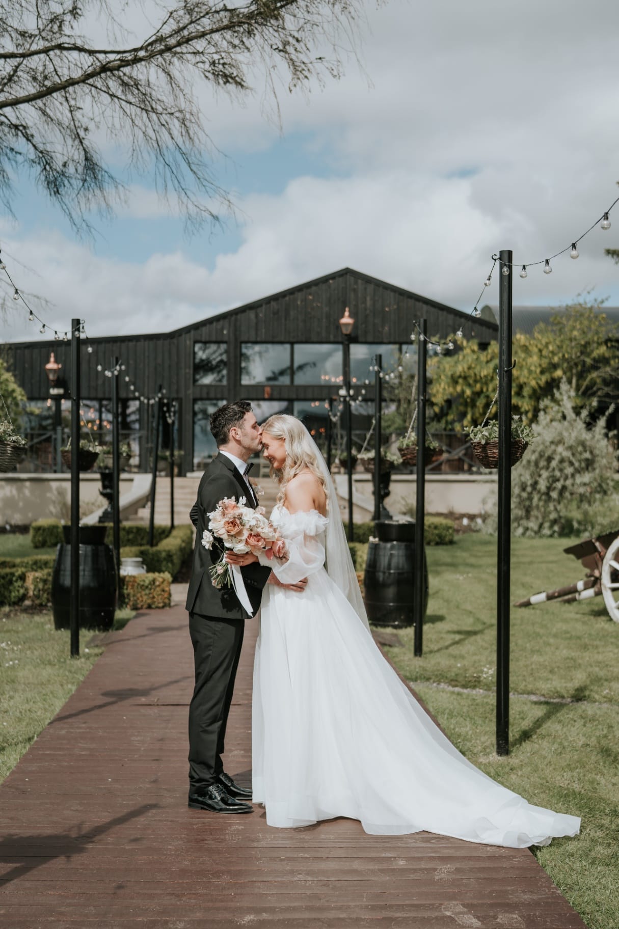 Groom kissing bride's forehead on the boardwalk outside The Barn