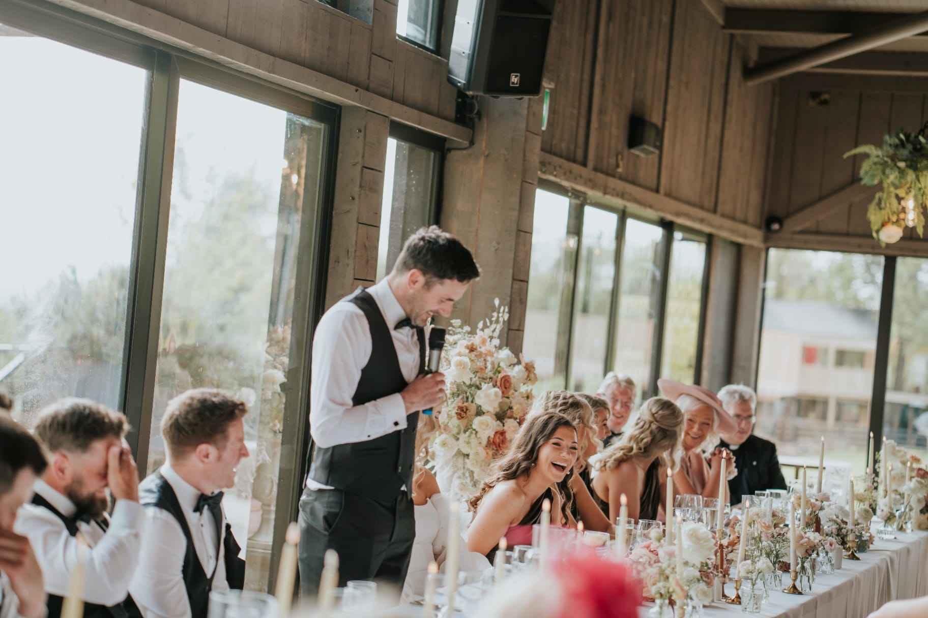 Groom delivering speech as bridal party laughs in sunlit barn reception